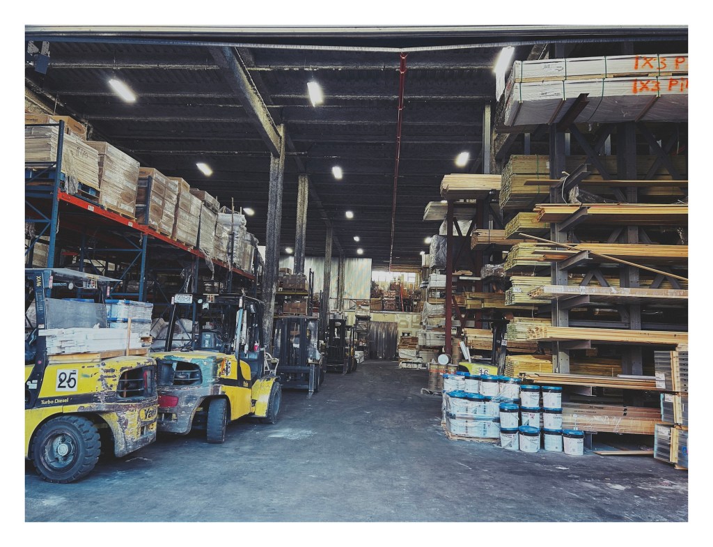 interior of a cavernous warehouse; tall shelves – filled with wooden boards and plants – line either side of a concrete pathway down the center of the frame