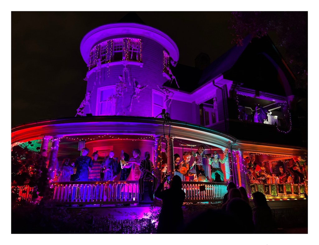 nighttime scene of a large Victorian home covered with Halloween decorations – skeletons, statues, animatronic monsters – and glowing with dramatic purple and orange lighting