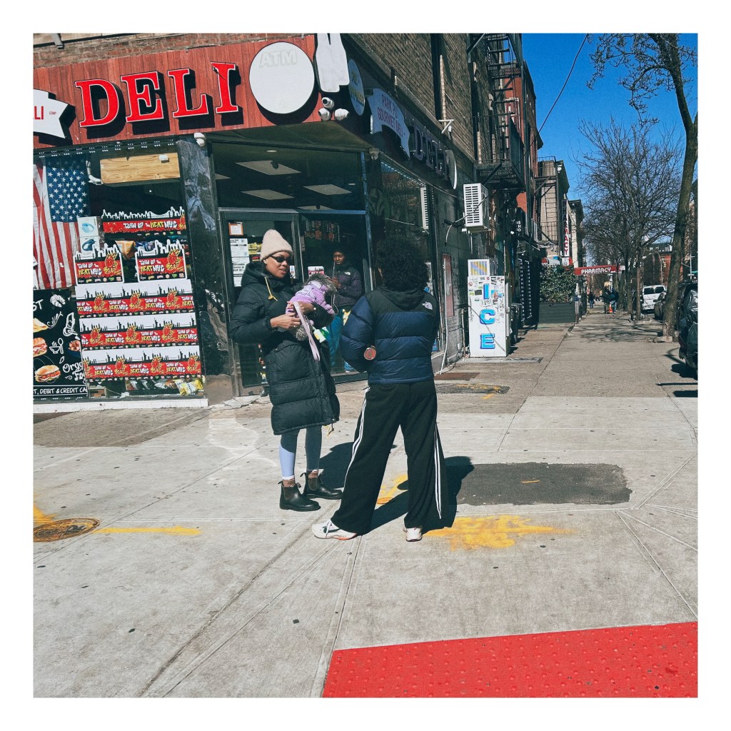 Two women stand on a street corner outside of a deli. One holds a small dog.