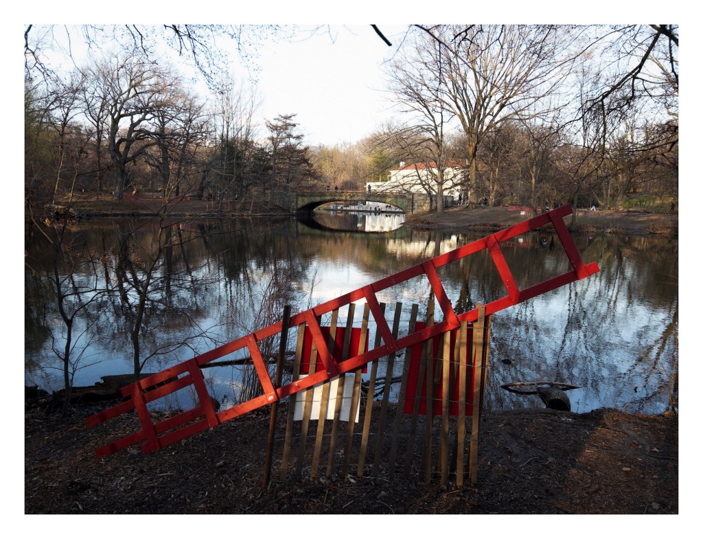 a red rescue ladder rests propped against a wooden fence alongside a pond; a bridge and boathouse are on the far side of the pond