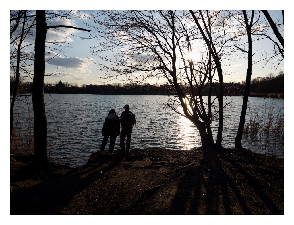 a woman and man scene from behind as they stand by the shore of a lake; the are silhouetted by the sun setting behind the far shore