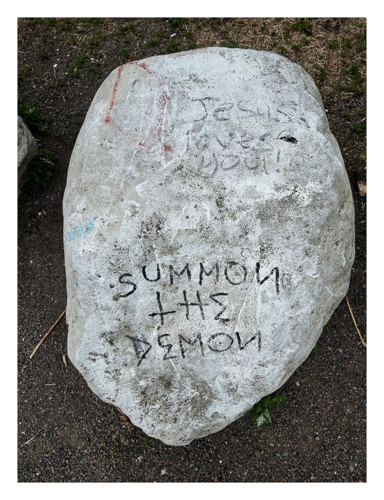 a pale boulder with two prominent graffiti writings: "Jesus loves you!" and "summon the demon"