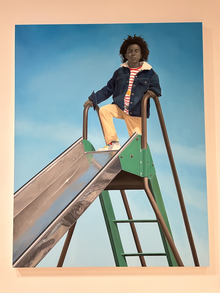 painting by Amy Sherald of a young boy in a denim jacket at the top of a slide with a bright blue sky behind him