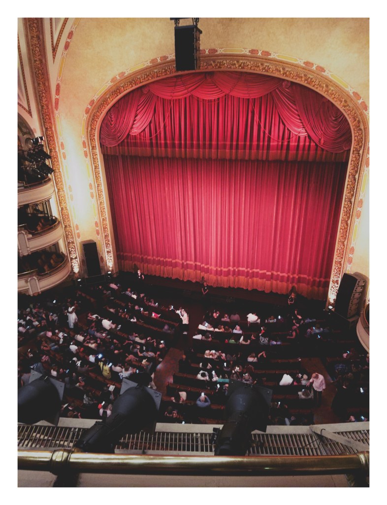 a view from the balcony of an ornate opera house, looking down toward the audience taking their seats in front of the main stage, closed off by tall red curtains