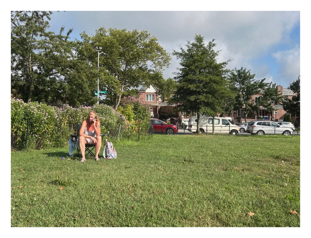a woman seated in a picnic chair on a lawn talks on her cellphone, with tall flowers and trees growing behind