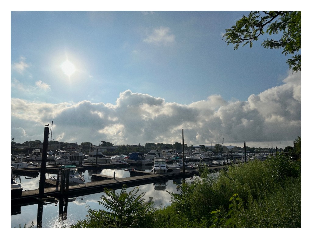 view of boats tied to wooden docks, with fluffy clouds and bright sun in the background