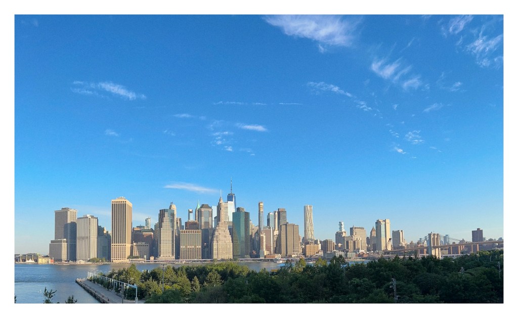 next to a river, tall skyscrapers buildings pierce a big blue sky; view of lower Manhattan skyline from Brooklyn Promenade
