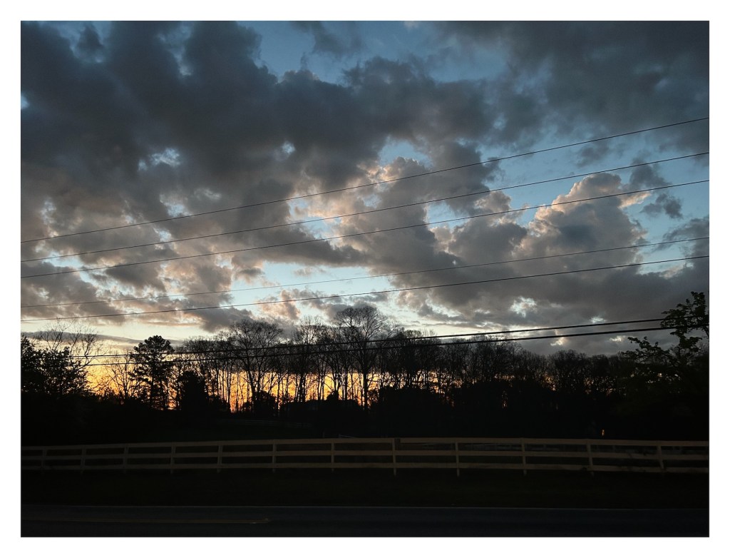 sunrise silhouettes a distant forest and lights the edges of clouds above a country road lined by a fence